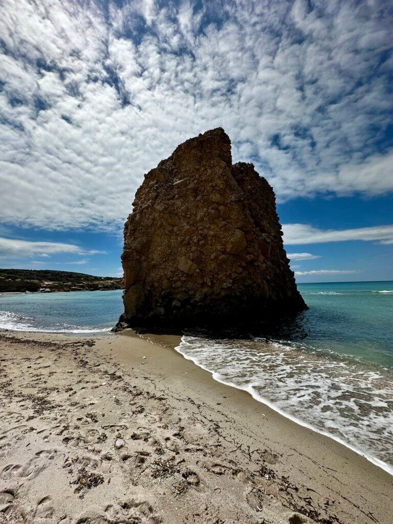 A solitary volcanic rock rising from a sandy beach in Milos beneath a textured sky